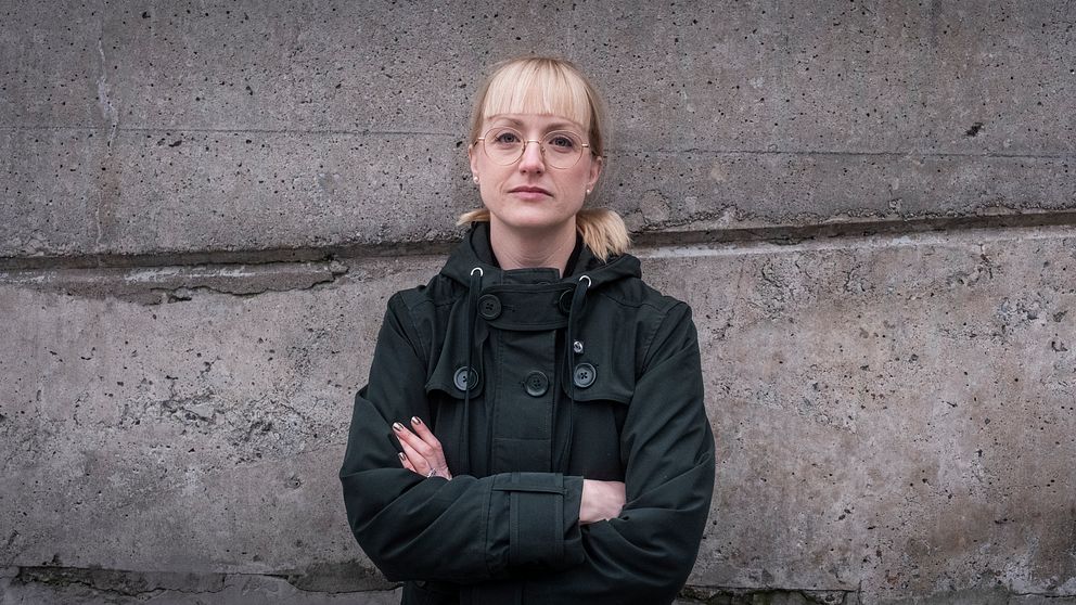 En person står med armarna i kors framför en sliten och fläckad stenvägg. Personen bär en mörk jacka och har sitt ljusa hår uppsatt i en knut.