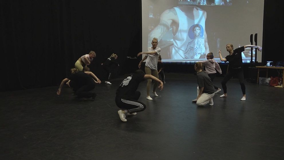 Grupp av dansare från sommardrömjobb projektet som övar på en scen, med en stor projektor i bakgrunden som visar en bild av en person. Dansarna utför olika rörelser, några sitter på golvet medan andra står och rör sig. Lokalen har mörka väggar och en enkel, svart golvbeläggning.