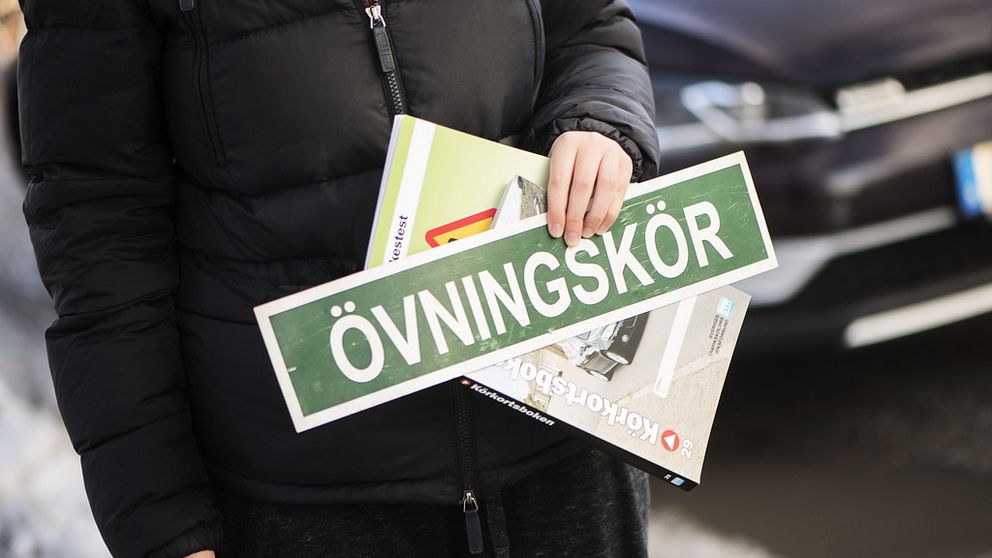 En anonym kvinna håller i skylten Övningskör samt en körkortsbok.