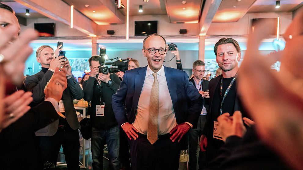 En grupp journalister och fotografer omger Friedrich Merz (CDU) i kostym under ett evenemang inomhus, och flera personer applåderar i förgrunden. Ljuset och inredningen antyder en professionell och festliknande atmosfär.