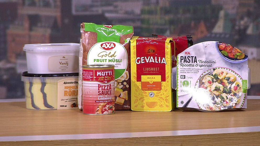Produkter från olika livsmedelsmärken står uppställda på ett bord. Från vänster till höger syns en burk med vaniljkräm, en förpackning med frukostmusli från AXA, en burk pizzasås från Mutti, en förpackning kaffe från Gevalia och en färdigpastarätt. Bakgrunden består av en suddig stadsbild.