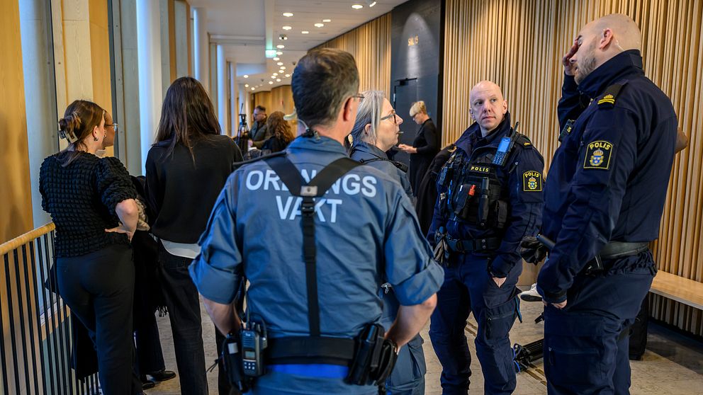 Poliser och bevakningspersonal står i en korridor och diskuterar, medan flera personer i civila kläder observerar situationen. Bakgrunden visar en modern inomhusmiljö med träpanel och bänkar.