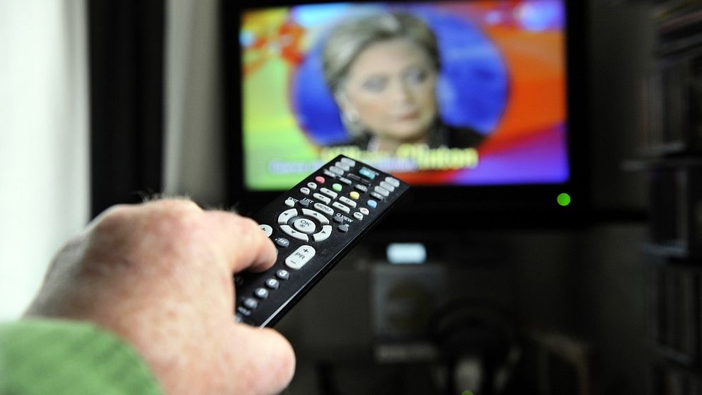 Bilden beskriver en man som håller i en fjärrkontroll. I bakgrunden på skärmen syns dem amerikanska politikern Hilary Clinton. Artikeln handlar om illegalt tv-tittande.