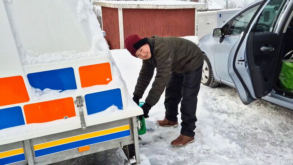 Alidad Zahedy lutar sig över en snötäckt släpvagn och fyller en flaska med vatten från kranen. I bakgrunden syns en parkerad bil och en röd träbyggnad, medan marken är täckt av snö.
