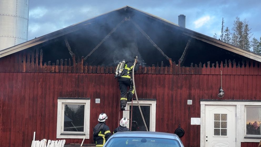 Brandmän arbetar på en röd byggnad i Avaträsk där rök stiger från taket. En brandman klättrar uppför en stege för att nå det rökfyllda området, medan en annan brandman står nedanför och övervakar situationen.