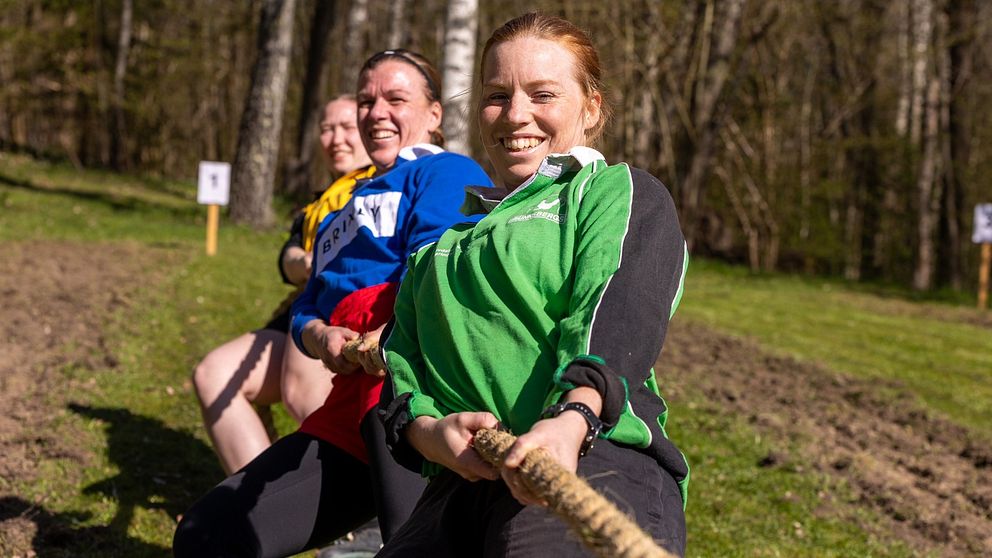 Tre personer drar i ett rep utomhus under en dragkamp i naturmiljö.