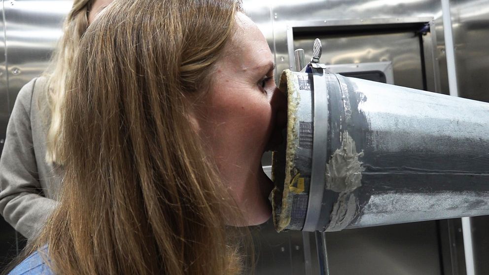 En person sitter bakom en megafonliknande apparat som ser ut att vara en del av en experimentell eller teknisk installation. Apparaten är grå med detaljer av silver och syns tydligt i förgrunden, medan en annan person står i bakgrunden och observerar.