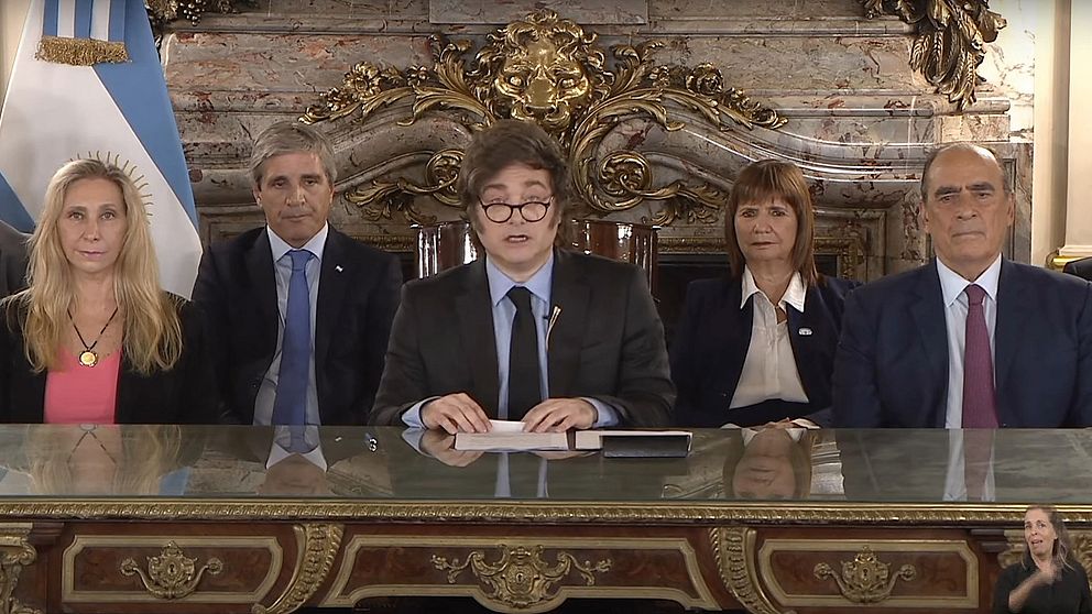 Fem personer i formellt möte vid ett dekorativt bord med argentinska flaggan i bakgrunden.