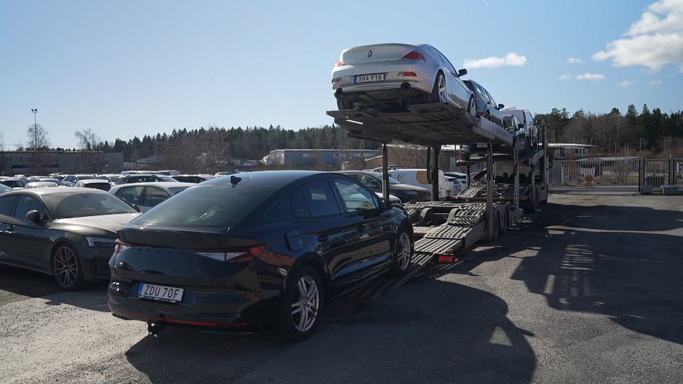 Bilar på en trailer vid en parkeringsplats under solig himmel.