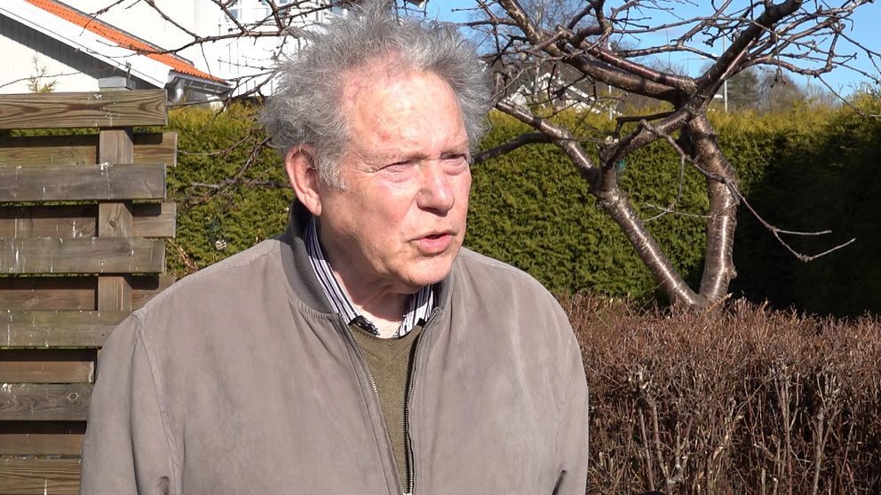 En person i neutral jacka står utomhus med buskar och ett träd i bakgrunden.