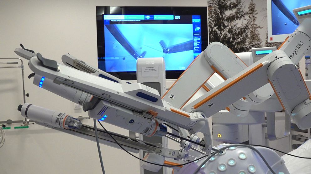 Robotassisterad kirurgiutrustning med flera rörliga delar i förgrunden, och en skärm i bakgrunden som visar en kirurgisk procedur. Utrustningen har blåa lysdioder och är placerad i ett modernt operationsrum.