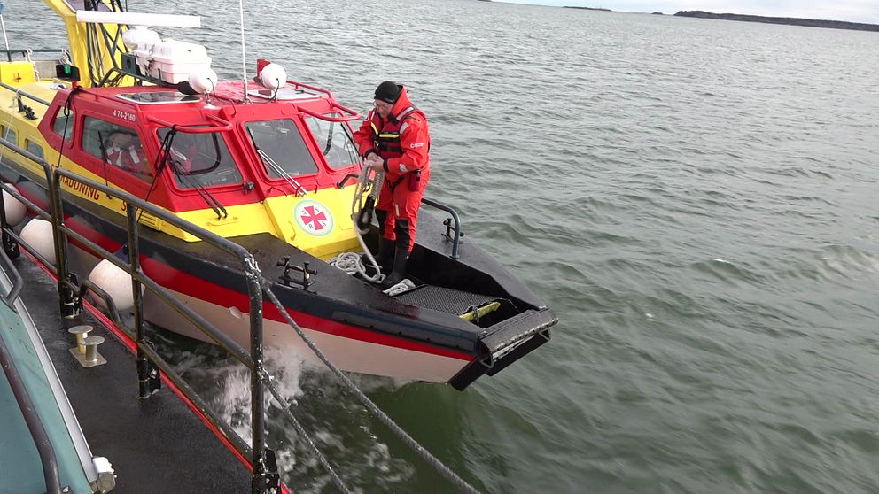 En räddningsbåt med röd och gul färg närmar sig en annan båt på vattnet. En medlem av räddningsteamet iförd röd utrustning förbereder sig för att gå ombord. Omgivningen visar lugna vatten och en klar himmel.