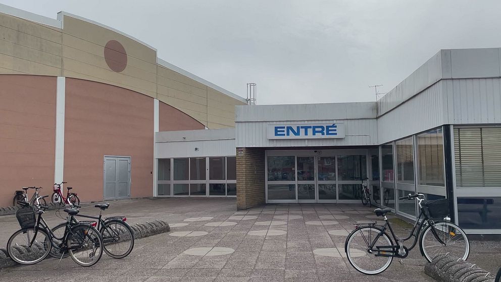 Entrén till en byggnad med glasdörrar, text ”ENTRÉ” ovanför. Två cyklar står parkerade på varsin sida om ingången på en öde stenlagd plats.