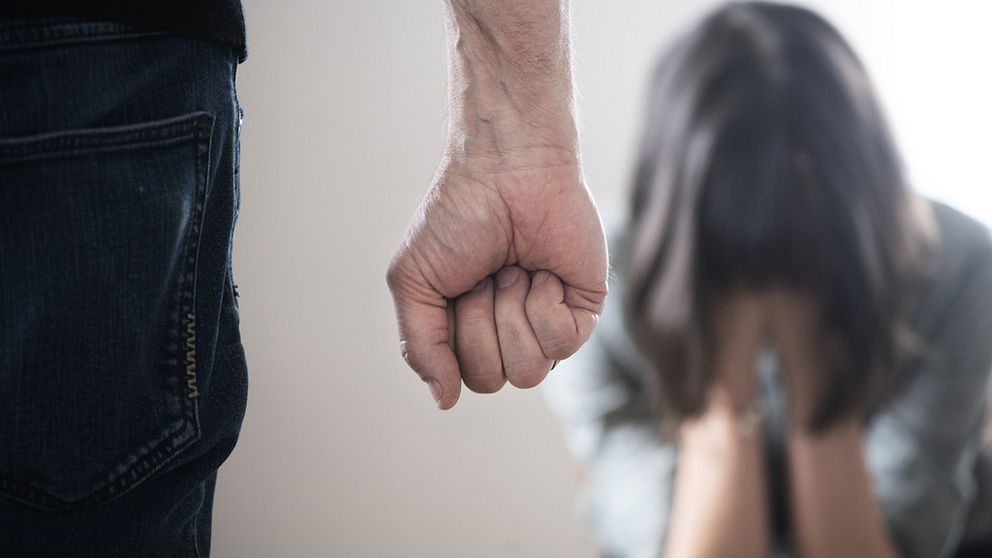 En knuten hand i förgrunden och en gråtande kvinna med brunt hår i bakgrunden.