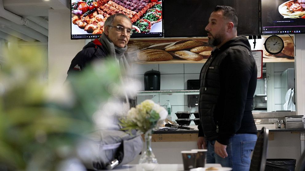 Två personer står och pratar i en restaurangmiljö. I bakgrunden syns en digital meny på skärmar som visar olika grillrätter. En blombukett och kaffemugg står på ett bord i förgrunden.