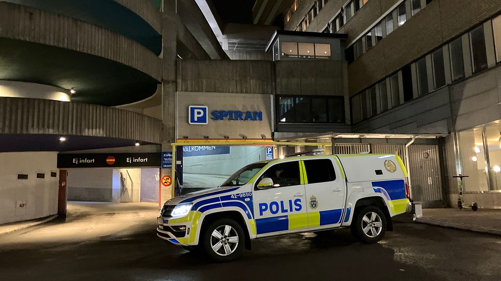 Polisbil parkerad vid ingången till ett parkeringshus med skylten ”SPIRAN” synlig i bakgrunden, omgiven av moderna byggnader i nattbelysning.