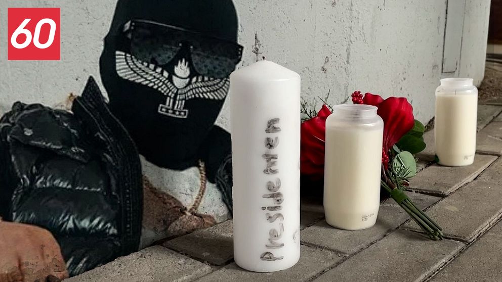 En man med en svart mask och solglasögon är klistrad mot en vägg. Framför honom finns två vita ljus och en röd ros placerade på marken. Denna scen kan tolkas som en hyllning eller minnesstund för en avliden person.