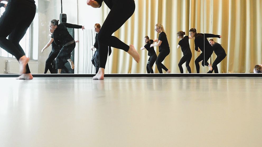 Flera dansare i svarta kläder utför en gruppdans i en studio med stora fönster och gula gardiner. Reflexioner i spegeln förtydligar rörelserna, medan fokus ligger på dansarnas fötter som rör sig i takt.
