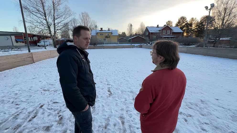 Två personer står i en snötäckt miljö med en byggnad i bakgrunden. Den ena personen är klädd i en svart jacka och den andra i en röd tröja. Trädkantade områden omger platsen och det är en klar himmel bakom dem.
