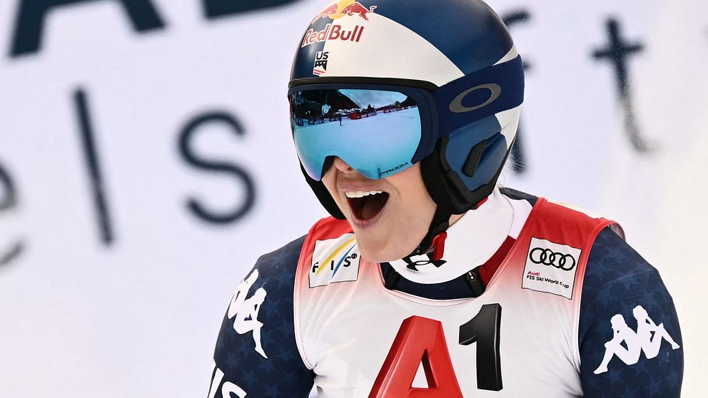 En alpin skidåkare i tävlingsdräkt med sponsormärken, förbereder sig för en nedfart. Skidåkaren har på sig en hjälm och en röd nummerlapp med bokstaven ”A1”. I bakgrunden syns en del av en stor reklamskylt med text.