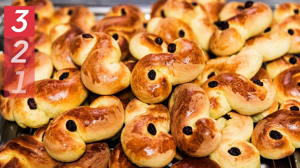 En stor mängd nybakade lussekatter, en traditionell svensk julbakelse, ligger på ett bakplåt. Bullarna har en gyllene yta och är dekorerade med russin. I vänstra hörnet finns en röd ruta med siffrorna 3, 2 och 1.