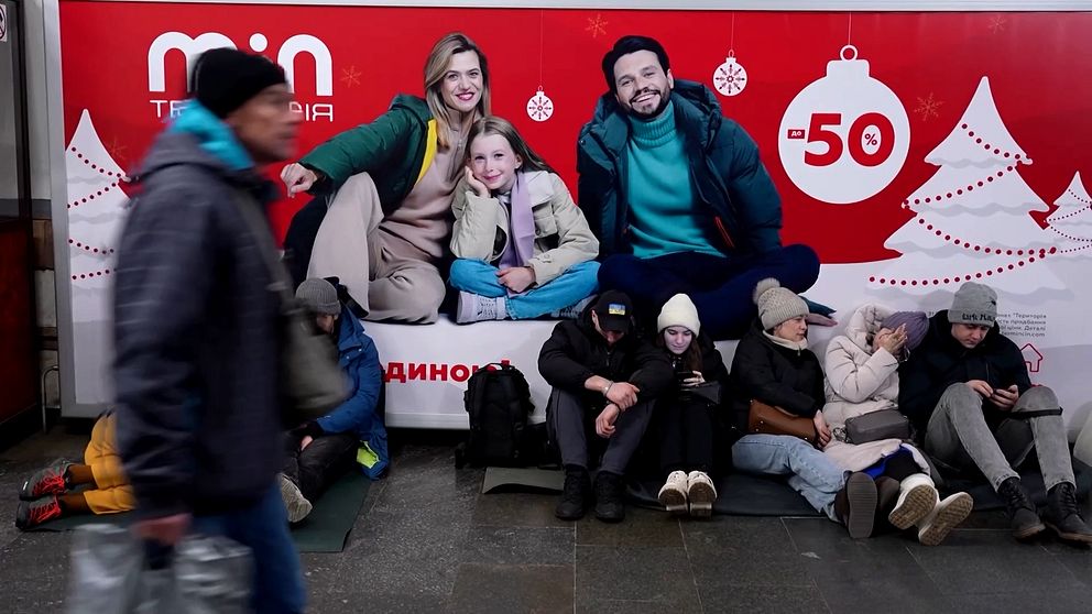 En grupp personer sitter på golvet i en tunnelbanestation, med en stor julskylt i bakgrunden som annonserar rea. Skylten är dekorerad med snöflingor och julgranartade motiv. Människorna är klädda i vinterkläder och några använder mobiler.