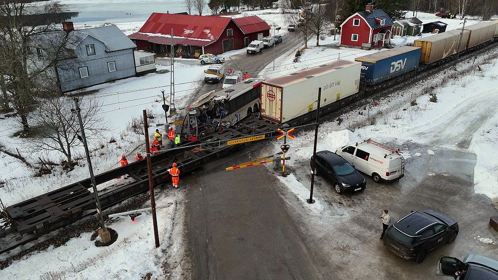 Fotot är taget från ovan och visar räddningspersonal som arbetar vid en järnvägskorsning i snöigt landskap. En lång godsvagn blockerar korsningen, och det står flera bilar parkerade i närheten. I bakgrunden syns några villor och en stor röd fastighet. På andra sidan spåret syns den demolerade bussen.
