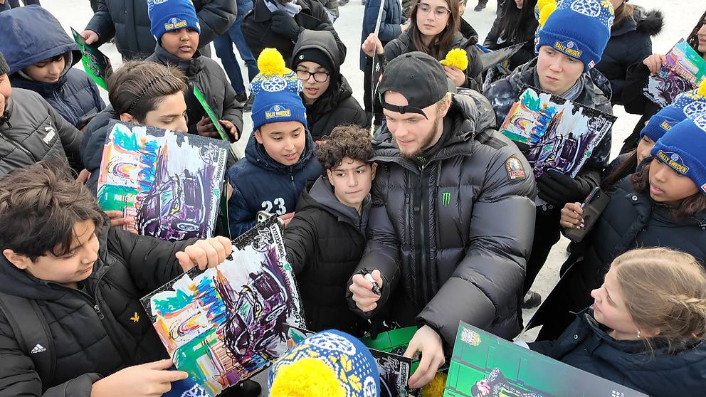 En grupp unga människor samlade runt en person som signerar autografer. De håller färgglada affischer och många av dem bär mössor och jackor som skydd mot kylan.