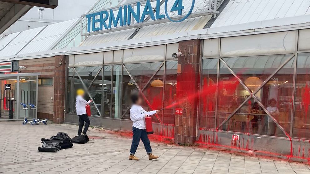 Två personer i vita kläder sprutar röd färg på glasfönstren vid Terminal 4. En av personerna står framåtlutad med en sprayflaska riktad mot väggen, medan den andra står till vänster med en svart väska på marken. Färgen droppar ner längs fönstren och markerar en vandalismhandling.