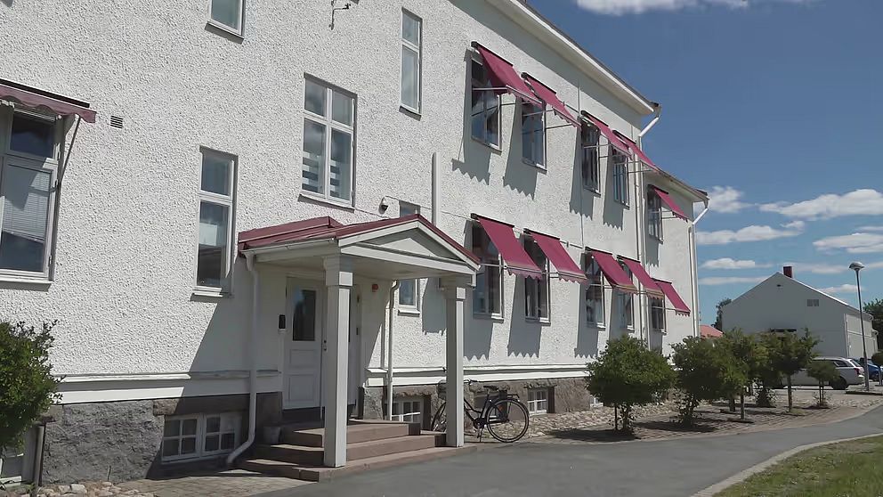 En vit byggnad med rödbruna markiser och en entré med tak. Det finns fönster på fasaden och en cykel stående på en sidogång. Grönska och buskar omger byggnaden, och i bakgrunden syns en annan byggnad och en blå himmel med några moln.