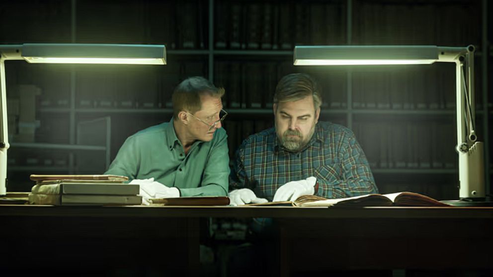 Två personer sitter vid ett bord i ett bibliotek, belysta av starka lampor, och bläddrar i gamla böcker. De har vita handskar på sig, vilket tyder på att de hanterar värdefulla eller ömtåliga volymer. I bakgrunden syns hyllor med fler böcker.