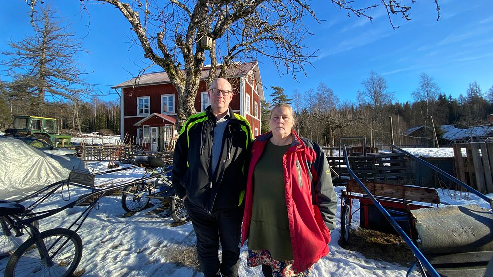 Man och kvinna står framför röd stuga i vintrigt landskap.