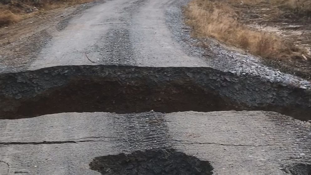 En grusväg med djupa hål och en stor spricka i mitten, omgiven av träd och buskar. Hålens kanter är ojämna och ytan ser skadad ut, vilket tyder på erosion eller annan typ av vägskada.