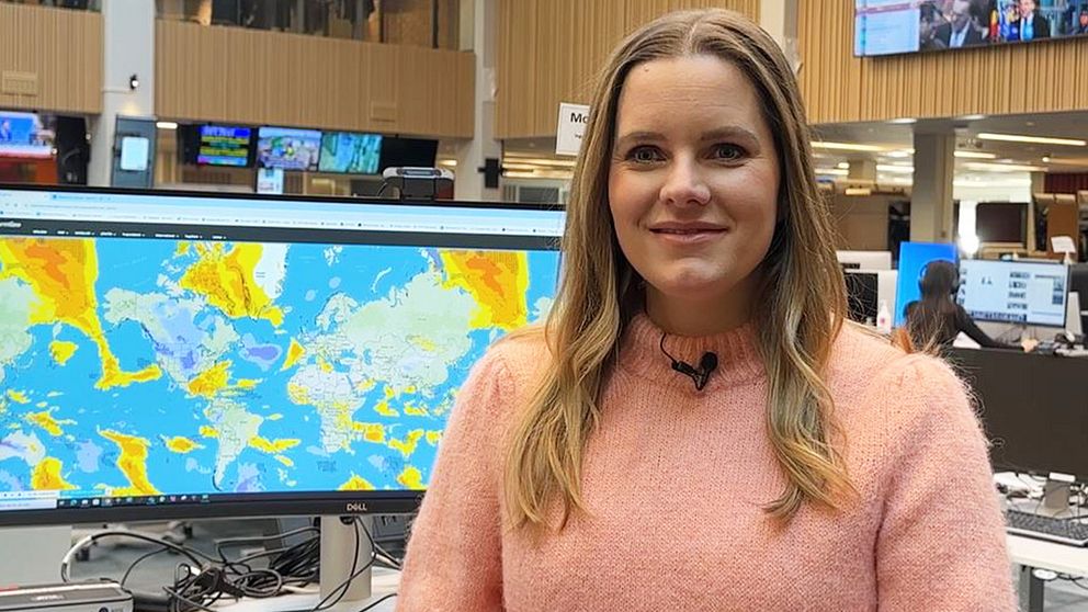 SVT:s meteorolog Tora Tomasdottir har en rosa tröja på sig och står framför en stor datorskärm i en nyhetsredaktion, med väderkartor synliga på skärmen. I bakgrunden syns fler skärmar och kontorsmiljö.