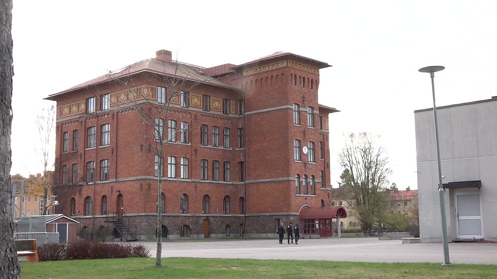 En stor röd tegelbyggnad med fyra våningar, som har detaljer som fönsterband och en klocka på väggen. Framför byggnaden syns en öppen yta med träd och några personer som går. En modern byggnad i grått syns till höger i bilden.