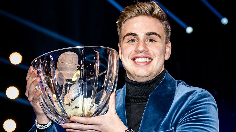 Personen i en blå sammetjacka håller upp en stor, klar glasbowl med en trofé inuti. Bakgrunden har starka ljusstrålar som skapar en festlig stämning.