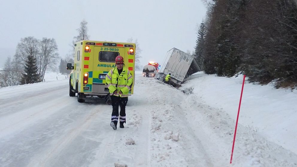 En lastbil ligger i diket intill en snöig väg. Framför står en ambulans och en man med gul väst kommer gåendes mot kameran.