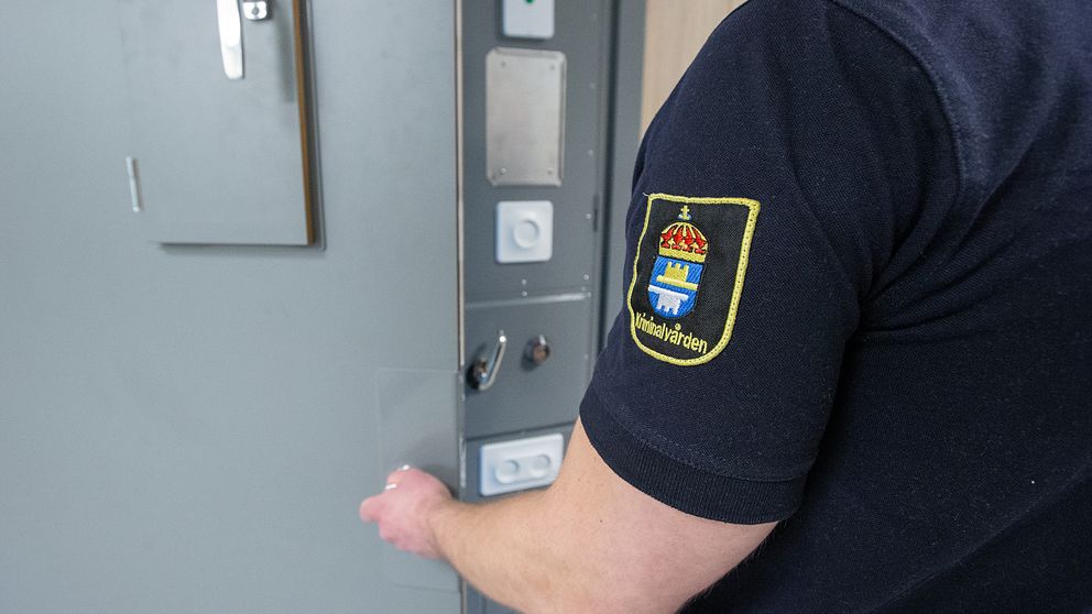 En person i en uniform med en emblem på ärmen öppnar en säkerhetsdörr. Dörrens panel har olika knappar och låsanordningar. Bakgrunden visar en neutral inomhusmiljö.