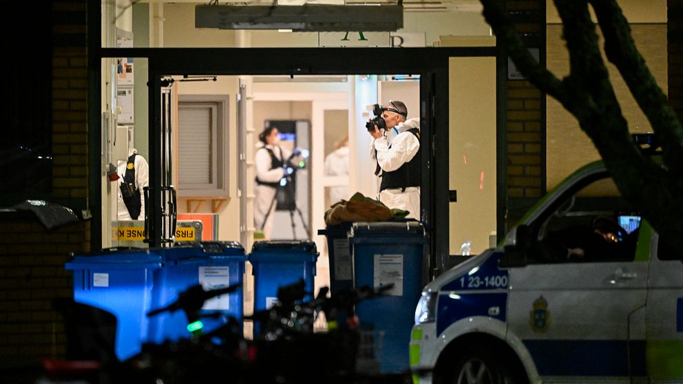 Kriminaltekniker arbetar inne i en byggnad, fotograferar och undersöker platsen. I förgrunden står en polisbil parkerad och flera soptunnor syns vid ingången.