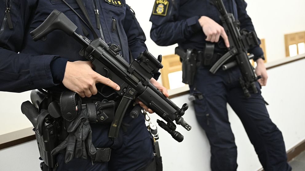 Två beväpnade svenska poliser i uniform håller i sina vapen inomhus, med fokus på deras utrustning och uniformmärken.