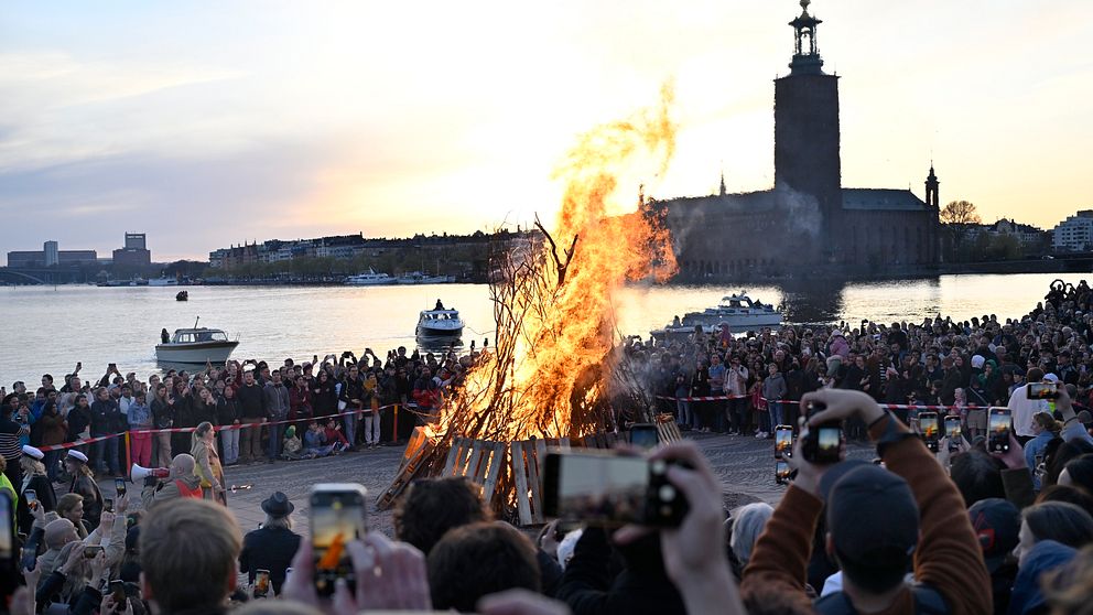 Stor folkmassa vid en majbrasa vid vattnet med Stadshuset i bakgrunden.