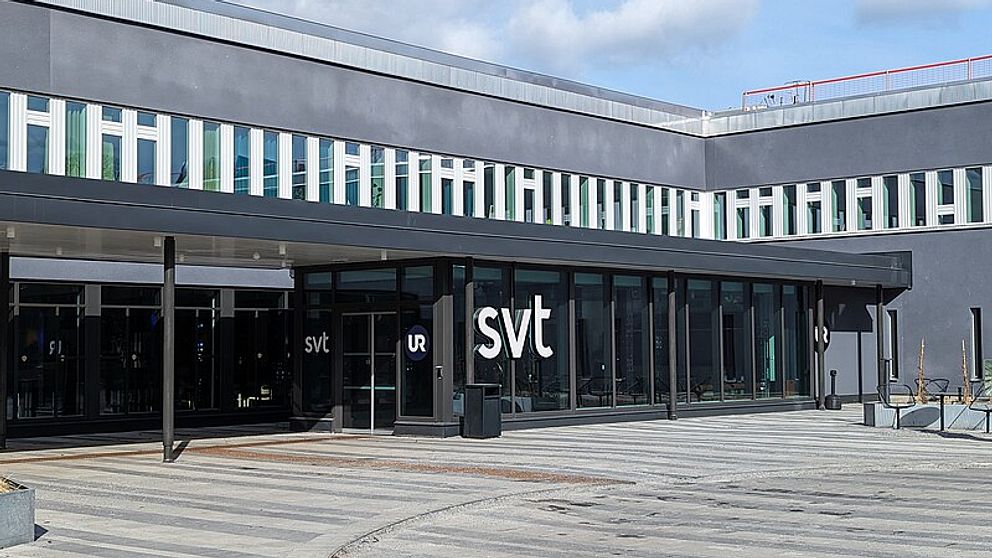 Entrén till SVT:s huvudkontor, med stora fönster och en modern fasad. Logotypen ”svt” syns tydligt på byggnadens framsida.