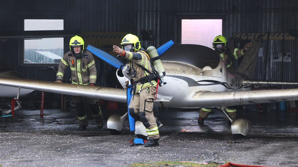 Brandmän i skyddsutrustning arbetar tillsammans med en liten flygplansmodell i ett hangar, med fokus på att säkerställa säkerheten och kontrollera propellern.