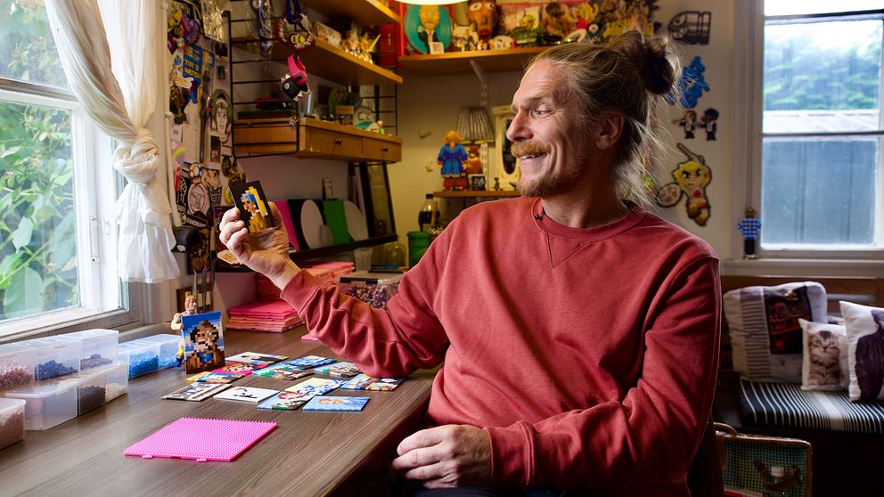 En person sitter vid ett bord i en kreativ arbetsmiljö, omgiven av färgglada dekorationer och konstverk på väggarna. På bordet ligger en uppsättning bilder och en rosa pärlplatta. Personen håller upp en av bilderna.