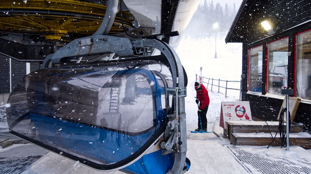 En liftkorg hänger från en skidlift i snöfall i Duved, medan en liftvärd sopar av påstigningsytan. I bakgrunden syns en skidbacke.
