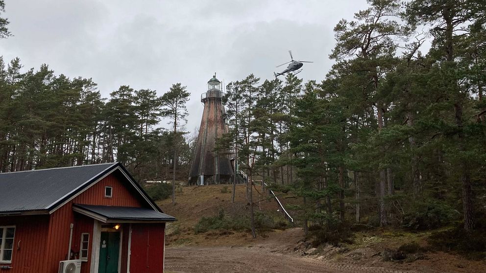 Röd stuga i skogsmiljö, med en fyr i bakgrunden på en mulen dag.