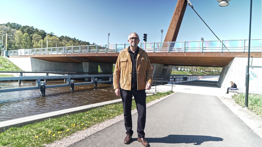 Man i brun jacka står på gångväg framför träbro vid kanal en solig dag.