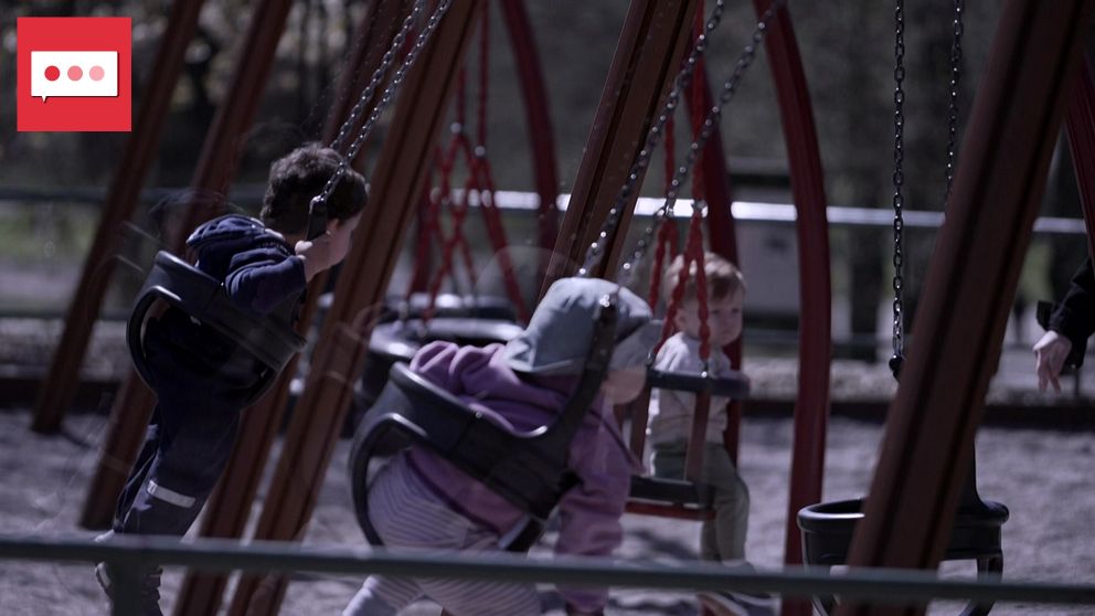 Tre barn leker på en gungställning i en park. Två av dem gungar, en pojke i en svart tröja och en flicka i gråa kläder, medan ett tredje barn sitter stilla på en gunga, klädd i ljusgrå kläder. Bakgrunden visar en grönskande miljö med träd.