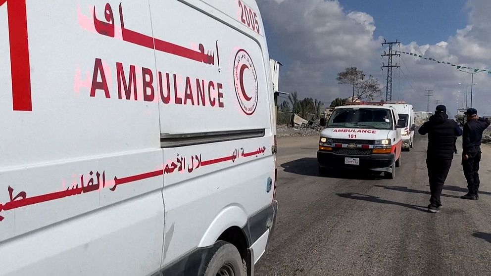 Två ambulanser står på en väg med tydliga röda kors och arabiska bokstäver på sidorna. Två personer i uniformer går bredvid ambulanserna under en molnig himmel.