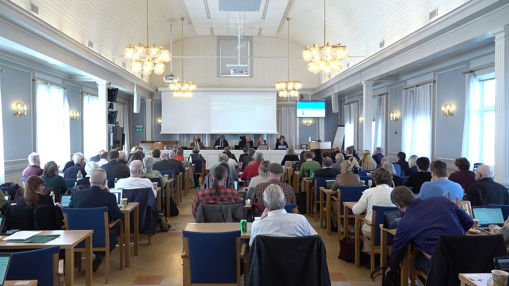 Möte pågår i stor konferenssal, deltagarna sitter vid bord och lyssnar på talare framme.